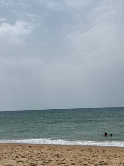 The image depicts a beach scene at Praia do Garrão, Almancil, Portugal, under an overcast sky. The foreground is composed of light brown sand, showing texture from footprints or natural undulations. A thin line of white foamy waves marks the immediate shoreline. The midground features the ocean, displaying a greenish-blue hue with visible surface ripples and small breaking waves. Two individuals are present in the water on the right side of the frame, partially submerged approximately to their chests. One individual appears to have dark hair or a dark swim cap, while the second, smaller individual is largely obscured. The background consists of a horizon line separating the ocean from a pale gray sky dominated by widespread, diffused cloud cover, with lighter patches indicating brighter areas. No other structures or objects are visible.