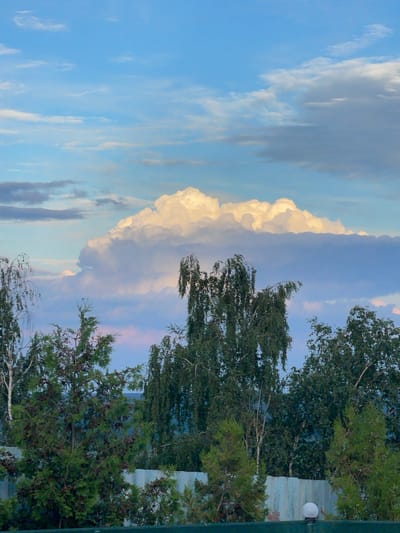 The image depicts a rural or suburban landscape under a cloudy sky, likely taken in Krasnokutsk, Ukraine. The foreground features a light blue fence with some greenery visible above it, including coniferous trees and larger deciduous trees. A single white lamp is attached to the fence on the right side. Above the trees, a massive, brightly lit cumulus cloud dominates the sky, suggesting either sunrise or sunset. The sky is a mix of blue and lighter, wispy clouds, with some darker, more distant clouds lower in the frame. There are no people or animals visible in the scene, and no actions or interactions are taking place.