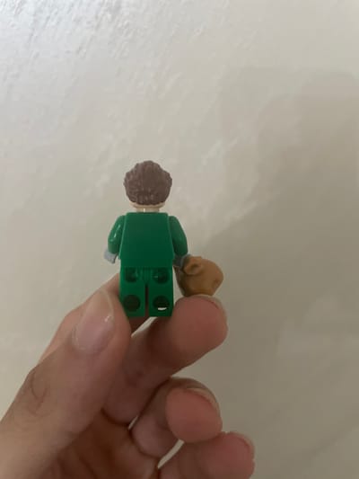 The scene, set in the city of Ihtiman, Bulgaria, depicts a human hand holding a Lego minifigure, viewed from the rear, against a plain, light-colored background. The minifigure has dark brown curly hair, a light nougat head, and is dressed in a dark green torso with light gray hands and dark green legs. Its right hand holds a small, tan-colored accessory. The details of the minifigure's back, including the leg connections, are clearly visible. The fingers of the hand holding the minifigure are visible, with neatly kept fingernails.