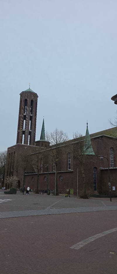 The image depicts the Sint-Lambertuskerk (St. Lambertus Church) in Horst, Netherlands, under an overcast sky. The church, constructed primarily of reddish-brown brick, features a prominent, tall bell tower on the left with arched openings, a visible clock face, an octagonal greenish-copper roof, and a golden cross finial. The main body of the church extends to the right, displaying multiple tall arched windows, smaller circular windows, and several additional greenish-copper spires and roof sections. Deciduous trees with bare branches are positioned along the church's façade.
In the foreground and mid-ground, a public plaza is visible, paved with a mix of reddish-brown and grey interlocking pavers. Two individuals are present on the paved area near the church's entrance on the left. One person, dressed in dark clothing, stands facing the church. The second person, wearing a white top and dark trousers, is seen pushing a white, wheeled object, possibly a stroller or walker. Several bicycles are parked or leaned against the church wall near public benches. A dark streetlamp is visible on the right. A small arrangement of flowers or a memorial is situated at the base of the church wall on the right side of the image. The overall lighting is diffuse, consistent with an overcast day.