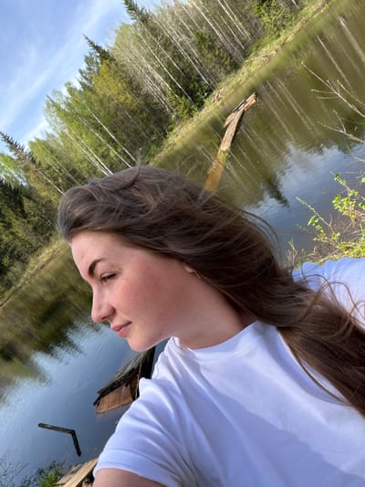 A young woman with brown hair and a white shirt looks down at a body of water. The water reflects the surrounding trees and sky. There is a piece of driftwood floating in the water. Ural'skoe, Russia is not evident in the image. The location cannot be confirmed without additional information.