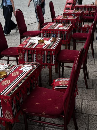 The image shows an outdoor seating area of a cafe or restaurant with tables and chairs set up on a cobblestone street. The tables are covered with red tablecloths featuring a black and white geometric pattern and red tassels. Several chairs are also visible, upholstered in a matching red fabric. Some tables have menus and ashtrays on them. In the background, a red Coca-Cola crate is visible. Two women are partially visible walking in the background, one wearing black pants and a blue top, and the other a patterned dress. The setting appears to be a pedestrian area in Sarajevo, Bosnia and Herzegovina, given the cobblestone pavement and outdoor cafe setup.