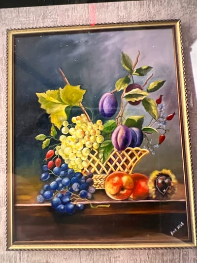 The image is a framed oil painting of a still life featuring a basket of fruit. The fruit includes grapes (both green and dark blue), plums, peaches, and what appears to be a chestnut. There are also some red berries and small blue flowers. The background is dark and atmospheric. The painting is signed in the bottom right corner with "Anna 2023". The frame is ornate and gold-colored, with a textured outer border. The painting is displayed in an indoor setting, with a textured wall visible behind it. There is no information provided within the image to indicate its location in the city of Bernex, Switzerland.