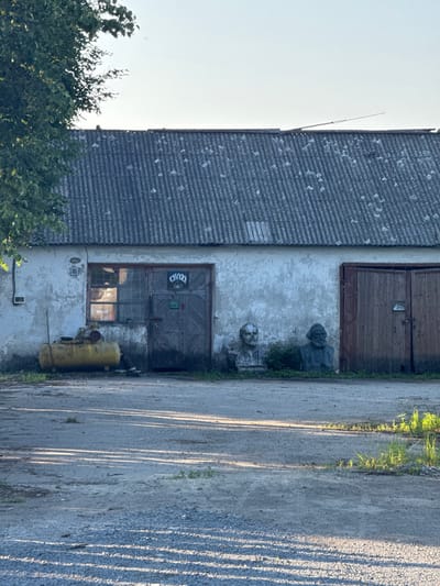 The image shows the exterior of a weathered, light-colored building with a corrugated metal roof. Two large, weathered wooden doors are visible on the right side of the building, and to their left is a smaller set of double wooden doors next to a window with multiple panes. In front of the building, to the left of the double doors, sits a large, horizontal yellow air compressor. Two busts, one of Lenin and another resembling Karl Marx, are placed on the ground in front of the building between the window and the right set of doors. Greenery and weeds grow around the bases of the busts and along the front of the building. The ground in front of the building is a gravel and dirt surface, with long shadows cast across it from an unseen light source. A large tree with dark green foliage is partially visible on the left side of the image, overhanging the building. The scene appears to be an outdoor yard or industrial area.
