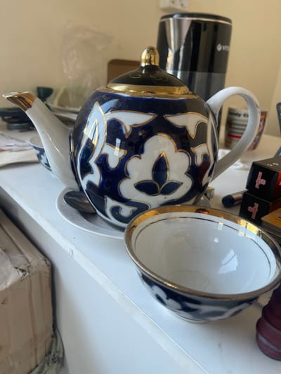 A close-up, eye-level shot captures a decorative teapot with a blue and white floral pattern, accented with gold trim, sitting on a white surface. A matching teacup, also with gold trim and a similar floral design, is placed in front of the teapot. The background is blurred, showing a black electric kettle and various other items on a shelf. The scene appears to be set in a home or an office, possibly Karasu, Uzbekistan, given the context. The lighting is natural, coming from the right, casting soft shadows on the objects.