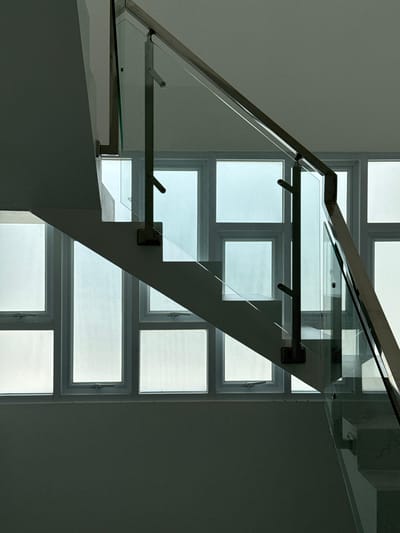 The image depicts a modern staircase with glass railings and a stainless steel handrail. The staircase is located indoors, as suggested by the windows and interior wall. The windows are large and frosted, allowing diffused natural light to enter the space. The overall aesthetic is clean and minimalist, with a focus on geometric shapes and materials. The color palette is neutral, consisting of whites, grays, and the cool blue-green tones of the frosted glass. The lighting creates a sense of depth and emphasizes the architectural lines of the staircase. There are no people or animals present in the image. The scene appears to be a residential or commercial interior, possibly a home or an office building. Without further context or identifying landmarks, it is impossible to confirm the specific location in Tanjungmeru, Indonesia.