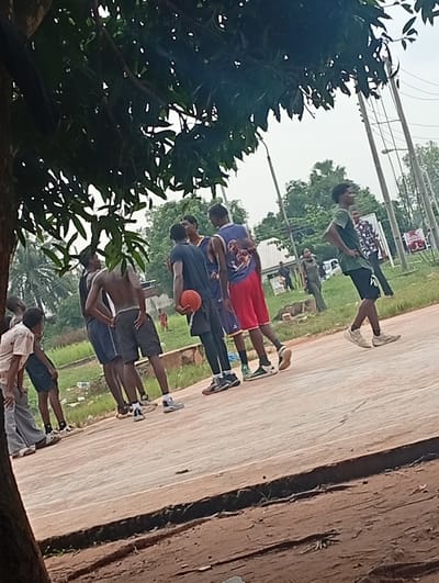 In Ukukwa, Nigeria, a group of young men are gathered on an outdoor basketball court, seemingly engaged in a game or practice. The court surface is a mix of concrete and dirt. The individuals are dressed in casual athletic wear, with some shirtless and others in jerseys and shorts. One person is holding a basketball, indicating an ongoing activity. The background reveals a lush green environment with trees and vegetation, characteristic of the Nigerian landscape. Utility poles and wires are visible in the distance, suggesting the presence of infrastructure within the community. The lighting suggests it is daytime, likely overcast or with diffused sunlight, as indicated by the soft shadows.