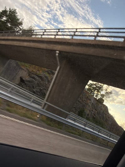 The image shows a view from a vehicle moving along a road, likely a highway, in Oskarsberg, Sweden. The perspective is from inside the vehicle, looking out and slightly upwards towards an overpass or bridge.
The overpass is constructed of concrete, with a solid lower section and a railing along the top. The railing has a distinctive pattern of rectangular openings, filled with a transparent or reflective material. Through the openings, a cloudy sky is visible, with a hint of trees and rocky terrain.
Beneath the overpass, a steep, rocky incline is visible, sparsely covered with vegetation. The road in the foreground is paved and has lane markings. A metal guardrail runs along the side of the road.
The lighting suggests it is either early morning or late afternoon, with the sun casting a warm glow on the sky and landscape. There are no people visible in the image. The overall impression is of a typical Swedish roadside landscape with infrastructure.