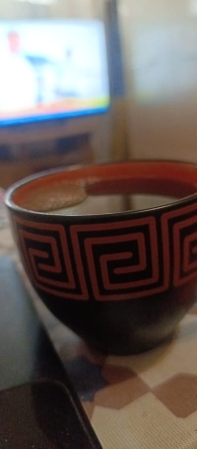 The image shows a close-up of a black and red patterned bowl filled with a dark liquid, likely tea or coffee. The bowl is resting on a patterned surface, possibly a tablecloth. In the blurred background, a television screen is visible, displaying a bright and colorful image. The scene appears to be indoors, in a casual setting. There are no people visible in the image. The location in the city of Voskresenskiy, Russia, is not discernible from the image content.