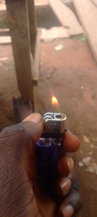 An individual's dark-skinned hand is holding a lit purple lighter. The lighter has a silver top with a ridged wheel and a flame issuing from its spout. The background is out of focus, showing a dirt ground and wooden structures. The scene appears to be outdoors in a rural or unpaved area, consistent with locations such as Baissa, Nigeria. The focus is on the lighter and the hand.