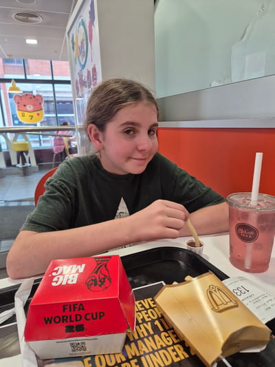 A young girl with brown hair sits at a table in a McDonald's restaurant in West Suffolk, United Kingdom. She is wearing a dark green t-shirt and is holding a french fry, about to dip it into a small cup of ketchup. In front of her are a red Big Mac box with FIFA World Cup branding, a box of fries, and a pink McCafe iced drink. The table is white and there is an orange wall behind the girl. In the background, other patrons and restaurant features are visible, including signage and seating.