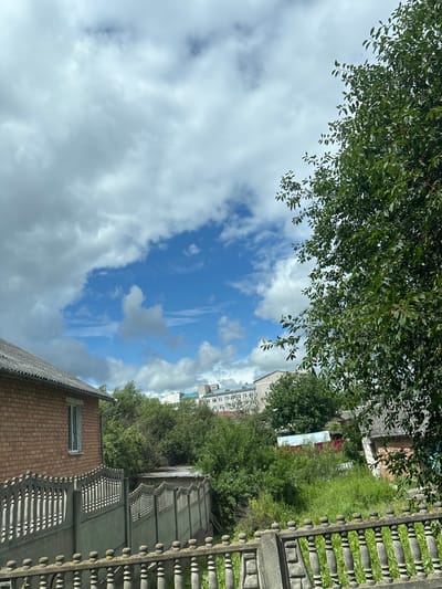 The image depicts a residential area under a partly cloudy sky in the Kapylsky Sel'sky Sovet, Belarus. In the foreground, a grey, ornate concrete fence runs horizontally across the bottom of the frame, with a section of a similar fence extending into the middle ground. To the left, a brick house with a grey roof and a single window is visible. Beyond the fence, dense green foliage and trees dominate the middle ground, obscuring much of the lower residential structures. In the background, a multi-story building with a light facade and numerous windows can be seen peeking through the trees. The sky is a mix of bright blue patches and large, fluffy white and grey clouds. A large tree with green leaves and some visible red berries is prominent on the right side of the image, extending into the upper right corner. The overall scene suggests a suburban or rural setting with evidence of residential development amidst natural greenery.