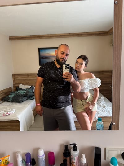 A man and a woman are taking a mirror selfie in what appears to be a hotel room in Ayvalık, Turkey. The man, with a beard, is wearing a black patterned polo shirt and grey shorts. He is holding a smartphone to take the picture. The woman, with dark hair pulled back, is wearing an off-the-shoulder white ruffled top and light yellow shorts. She is standing next to him, with her hand near her shorts. In the background, two beds are visible, one with patterned pillows and the other with white bedding. There is a framed picture on the wall above the beds, depicting a sunset over a body of water. A shelf below the mirror holds various toiletries and a bottle of water. The room has light-colored walls and wooden accents.
