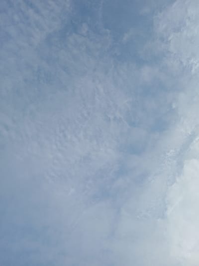 A bright sky, predominantly pale blue, fills the frame. Thin, scattered clouds create a textured effect. Towards the lower right, larger, whiter cloud formations appear, contrasting with the finer, more uniform clouds in the upper and left sections. The overall impression is airy and expansive.