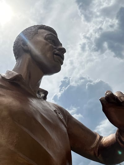 This is a low-angle shot of a bronze statue of two figures in a kissing embrace, set against a cloudy sky. The statue is positioned outdoors, with the sun peeking through the clouds, creating lens flare and highlighting the metallic sheen of the sculpture. The perspective is from below, looking up at the figures' faces and upper bodies. The sky is a mix of grey and white clouds, suggesting either overcast conditions or a dynamic, changing weather pattern.

The statue appears to be made of bronze, with a polished finish that reflects the light. The figures are depicted in a close embrace, with one figure's head tilted back and the other leaning in for a kiss. The details of their faces are somewhat softened by the material and the lighting, but their expressions convey a sense of intimacy and passion. The musculature of their bodies is also evident, suggesting athletic or strong builds. One figure has a small emblem or detail visible on their shoulder or upper arm.

The time of day appears to be daytime, given the presence of the sun and the brightness of the sky, though the cloud cover suggests it might be a partly cloudy day. The location is identified as Staryi Aidar, Ukraine, but there are no specific visual cues in the image to confirm this. The overall mood is romantic and possibly celebratory, given the public placement of a statue. There is no visible text in the image.