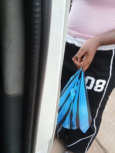 The image depicts a partial view of an individual in Orie Uratta, Nigeria, standing next to the open door frame of a vehicle. The person is wearing a light pink top and black athletic shorts or pants with a white elastic waistband and white vertical stripes. The numeral "08" is visible in white on the right leg of the garment. The individual's right hand, with fingers curled, grips the tied handles of two plastic bags: one light blue and one black. These bags contain an unidentifiable dark-colored item or items. The vehicle door frame, visible on the left, is white, while the interior panel behind it is dark grey. A light brown, unpaved ground surface is visible beneath the person.