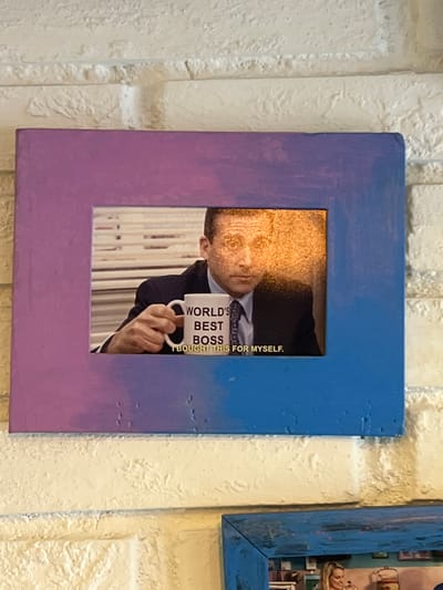 The image shows a framed picture of Michael Scott from The Office holding a mug that says "World's Best Boss, I bought this for myself." The frame is painted with a gradient from purple to blue. The framed picture is hanging on a white brick wall. Below it, another framed picture with a blue frame is visible. The overall scene appears to be an interior setting, possibly a home or an office space, located in Maracay, Venezuela. There are no people visible in the immediate foreground of the image, only the framed artwork.