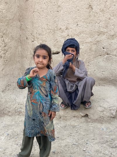 This media file features two young children in Arghandab, Afghanistan. The child in the foreground is a young girl wearing a long-sleeved dress with a blue, green, and brown floral print. She has dark hair and is looking towards the camera with a slight smile. She is adorned with several green bracelets on her left wrist. She appears to be standing on or near a dusty, uneven ground.

In the background, a slightly older child, possibly a boy, is seated. This child is wearing a grey tunic and trousers and a dark blue turban that covers their head and part of their face. Their hand is partially covering their mouth, and they are looking at the camera with a direct gaze. The background wall is rough and textured, made of a light-colored material, possibly mud or stucco, showing signs of wear and cracking. The ground is a mixture of dirt and small stones.

The lighting suggests it could be daytime, possibly late morning or afternoon, with diffused sunlight. The overall scene depicts a candid moment of two children in a simple, possibly rural, setting. The children's expressions are neutral to slightly curious or engaging with the photographer. There is no visible text in the media file.