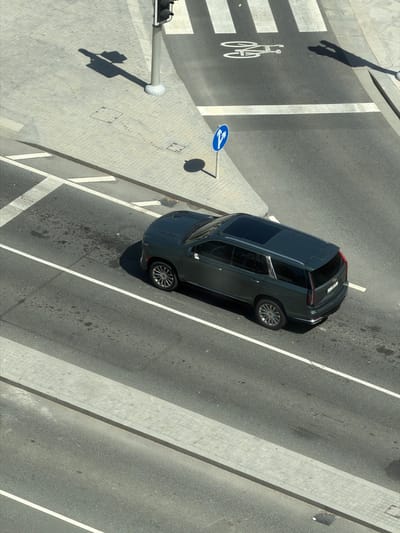 An overhead photograph captures a dark grey vehicle, likely a crossover or compact SUV, situated diagonally on an asphalt road in Lusail, Qatar. The car features a sunroof and multi-spoke wheels, positioned across a lane marked with white solid lines.
To the left of the vehicle, a white crosswalk is painted on the road, prominently featuring a white bicycle symbol, indicating a bicycle lane or crossing. Adjacent to this, a light grey paved sidewalk extends, showing part of a traffic light pole and another pole casting a long shadow. A distinct, elongated human-like shadow is cast across the crosswalk area.
Further down the road, a blue circular sign with two white arrows (one pointing left, one pointing right) is mounted on a pole, likely indicating permitted turns or a forthcoming roundabout. The scene is brightly illuminated by direct sunlight, creating sharp, elongated shadows from the car, poles, and the human figure, suggesting the time of day is either mid-morning or late afternoon. The paved sidewalk continues to the right side of the image, where a drain grate is visible. The car's rear license plate is present but its text is illegible.