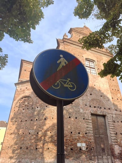 This image shows a traffic sign that indicates the end of a no-pedestrian and no-cycling zone. The sign is circular with a blue background and a red diagonal slash across it. Inside the circle, there is a white silhouette of a person walking and a white silhouette of a bicycle. The sign is mounted on a dark pole. In the background, there is a brick building with a large wooden door and a cross on top. The building appears to be a church or a historical structure. There are also some trees with green leaves visible in the foreground and background. The sky is blue with some clouds. The overall scene is an outdoor shot, likely in Italy, as indicated by the architecture and the traffic sign, which is common in Italian cities. The lighting suggests it is daytime.