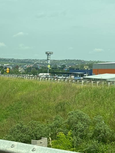 The image shows a wide shot of a commercial area on the outskirts of Solontsy, Russia. In the foreground, there is tall, green grass and some shrubbery. A concrete barrier with visible bolts runs along the bottom of the frame. In the middle ground, a highway or major road is visible with a white barrier separating it from the grassy area. Several cars and a truck are parked in a lot beyond the road, suggesting a commercial establishment. A large sign for "METRO" is prominently displayed on a tall pole, indicating a Metro Cash & Carry store. Adjacent to this, there is a large building with an orange and gray facade, and another building with a blue facade. In the background, a densely populated residential area with numerous houses and buildings stretches across rolling hills. The sky is overcast and a pale blue-green color. There are no people clearly visible in the image.