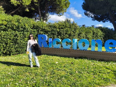 A female subject stands on a grassy lawn in front of a large, three-dimensional "Riccione" sign, located in Riccione, Italy. The sign, composed of individual letters in various shades of blue, is mounted on a low, dark-colored base. The subject, positioned to the left of the sign, wears a light-colored long-sleeved top, light blue baggy jeans, white sneakers, and sunglasses. Her dark hair is down. She holds a dark, fluffy item under her left arm and a small cluster of yellow items in her right hand. A distinct shadow from her body is cast on the grass to her left. Behind the sign and extending into the background are dense green bushes and several tall trees. The sky is clear blue with scattered white clouds. The ground is covered with green grass interspersed with small white flowers.