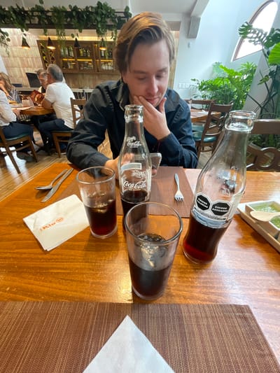 A young man with light brown hair is seated at a wooden table in what appears to be a restaurant. He is wearing a dark blue button-down shirt and is looking down at the table, with his right hand resting on his chin. In front of him are two glasses filled with a dark liquid and ice, and two glass bottles of Coca-Cola. One bottle is closer to the foreground and is partially visible, while the other is further back. A white napkin with an orange logo, cutlery (a spoon and a knife), and a fork are also on the table. In the background, other patrons are seated at tables, and a shelf unit with bottles is visible. Plants are arranged in the environment, and part of a slanted ceiling with a skylight is also present. The overall setting suggests an indoor dining establishment in Cuautitlán Izcalli, Mexico, based on the presence of the "Exceso Calorías" and "Exceso Azúcares" labels on the Coca-Cola bottle, which are common in Mexico.