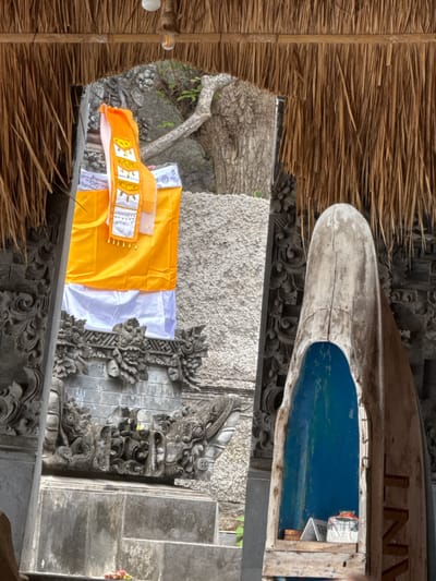 An outdoor, eye-level, medium shot captures a religious shrine in Abang, Indonesia, under a thatched roof. In the center, an orange and white fabric hangs on a carved stone pillar, a common offering at Indonesian temples. To the right, a weathered, boat-shaped wooden structure with a bright blue interior stands against another stone pillar. The shrine is made of grey stone, adorned with intricate carvings of mythical creatures. The background reveals lush green foliage and trees, suggesting a tropical setting. The lighting is natural and soft, indicating it was taken during the daytime.