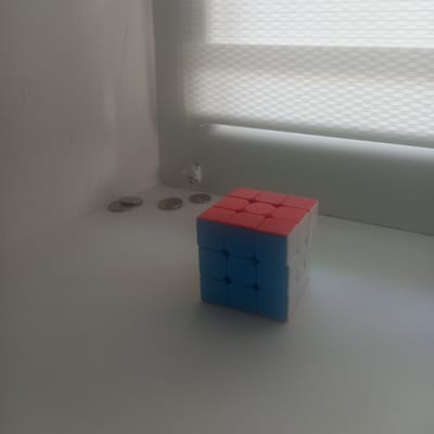 The image depicts a close-up, eye-level, slightly angled shot of a Rubik's Cube positioned on a flat white surface. The cube exhibits red and blue colored faces, with some white faces also visible. To the left of the cube, several silver coins are scattered on the same white surface. In the background, a window with horizontal blinds is visible, suggesting an indoor setting. The lighting appears to be natural, coming from the window, casting soft shadows. The overall environment suggests a quiet, possibly domestic setting. The scene is static and does not depict any activity or interaction. No people are present in the frame. The location in Cepeleuți, Moldova, is inferred from external context, but is not visually evident in the image itself.