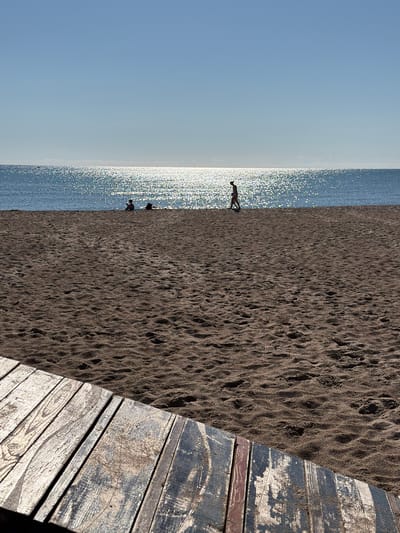 A full vertical shot captures a sunny day at a beach in Lozenets, Bulgaria. In the foreground, a weathered wooden boardwalk runs diagonally from the bottom left corner to the bottom right. The boardwalk planks are varied in color, showing signs of age and exposure to the elements, with wood grain, paint chips, and sand evident. Beyond the boardwalk, the sand-covered beach stretches towards the shimmering, sun-drenched sea. The water is a deep blue, reflecting the bright sunlight, creating a sparkling effect across its surface. In the distance, along the shoreline, three figures are visible. One figure, a silhouette, is walking along the water's edge from left to right. To the left of this walking figure, two other figures are seated or lying on the sand near the water. The sky above is a clear, pale blue, devoid of clouds. The overall scene depicts a peaceful, summery beach atmosphere.