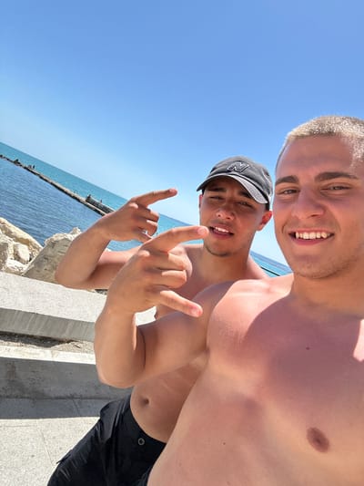 The image shows two young men posing for a selfie outdoors, likely at a beach or seaside location in Balchik, Bulgaria. The man in the foreground, wearing a black baseball cap, smiles broadly, his mouth open as if mid-sentence or laughing. He holds up his hands in a rock-and-roll gesture. Behind him, another man, with short blond hair, also smiles at the camera, his face partially obscured by the angle of the shot. Both men are shirtless, suggesting warm, sunny weather.
The background features a bright blue sky and the calm, glistening sea. A concrete structure, possibly a pier or seawall, runs along the edge of the water, with some rocks visible near the foreground. The sun is high in the sky, indicated by the strong lighting and shadows. The overall mood is one of fun and camaraderie, with the men appearing relaxed and happy. The lighting suggests it is daytime, likely midday, given the brightness of the sun. No visible text is present in the image.