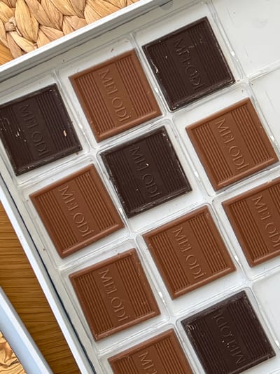 The image shows a close-up, overhead view of an open box of chocolates. The box is white with a clear plastic divider holding twelve individual chocolates. Nine of the chocolates are visible, arranged in three rows of three, with the bottom right corner of the box cut off. Each chocolate is rectangular with a ridged surface and embossed lettering that reads "MELODI". There are three different shades of chocolate visible: dark brown, medium brown, and a lighter milk chocolate. The box is placed on a textured surface, possibly a woven placemat, with a light brown and straw color. The lighting is bright, creating some glare on the chocolates. There are no people or actions depicted. The image appears to be a product shot or a personal photo of the chocolates. There is no information available to indicate the location in the city of Şişli, Turkey.