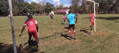 A soccer game is taking place on a field with green grass. There is a soccer goal in the foreground and several players are visible in the background, all of whom are wearing red or blue jerseys. The player in the foreground, facing the goal, has a red jersey with the number "5" on the back. The player is running toward the goal,  holding the soccer ball. The coordinates 0.554, 34.773 place this location in Bungoma, Kenya.  The image was captured in daylight.  A small building can be seen in the background of the image, and there are trees in the background as well.  There are other people watching the game in the background.
