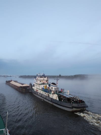 A tugboat, identified by the lettering "OTA-922" on its stern, tows a barge loaded with light-colored aggregate down a river. The tugboat has a white superstructure with several decks, and a dark hull. The barge is long and flat, with the aggregate piled high in the center. The river is calm, with a slight chop on the surface. Mist hangs low over the water and the distant tree-lined shore, suggesting an early morning or late evening setting. The sky is a pale blue with some scattered clouds. The scene appears to be on the Volga River near Chaika, Russia, a town known for its river port and industrial activity. No people are visible in the image.
