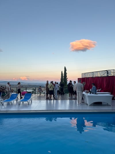 A group of people are gathered on a deck next to a swimming pool, with a scenic view of hills and a town in the distance. The sky is a pale blue with a single, fluffy orange cloud. The sun appears to be setting, casting a warm glow across the landscape. Some people are standing in a line, seemingly waiting for something, while others are seated on lounge chairs. There's a table with what appears to be a buffet or bar setup, complete with red drapes and white coverings. The overall atmosphere appears to be relaxed and social, possibly at a party or event. The image is oriented vertically.