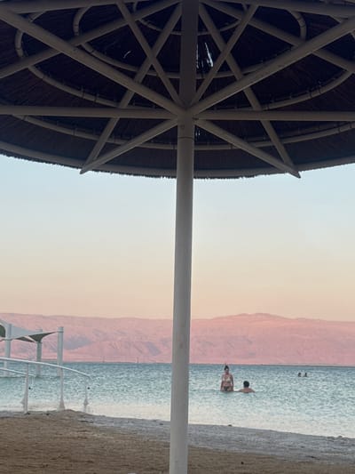 The image depicts a scene at the Dead Sea in Tamar Regional Council, Israel. In the foreground, a portion of a thatched umbrella structure is visible, with its white metal frame extending across the left side of the frame. The umbrella provides shade, suggesting a sunny day.
In the middle ground, the Dead Sea is observed with people enjoying the water. Two individuals are visible wading in the water, with one appearing to be partially submerged. In the background, a range of reddish-brown mountains forms the distant landscape, characteristic of the Dead Sea region. The sky is a pale, hazy gradient, indicating either early morning or late afternoon light, with a soft glow.
The overall atmosphere appears relaxed and recreational, with the individuals in the water seemingly at leisure. The setting is a beach or shoreline, with the sand visible on the right side of the frame. No visible text is present in the image.