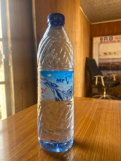 A plastic water bottle, branded "Mr. V Premium Water" with a blue cap and a blue and white label indicating "75cl," stands upright on a polished light brown wooden surface. The bottle contains clear liquid and has a textured, ribbed design. In the blurred background to the left, a bright light source, likely a window, is visible. To the right, also blurred, an indoor environment with wood-paneled walls features a dark office chair with chrome accents and a framed poster or picture displaying "DRB" at the top. The ceiling appears textured. The specific location within Gyellesu, Nigeria, is not discernible from the visual information provided. No individuals are present, and no actions or interactions are depicted.