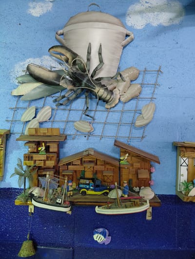 The image displays a nautical-themed wall decoration, likely within a home or establishment in Cutler Bay, Florida. The scene presents a blend of sculpted and crafted elements mounted on a blue-painted wall.
On the left, a large, sculptural representation of a lobster emerges from a pot. This assembly is connected to a wire mesh, supporting what appear to be stylized shell shapes.
Moving to the right, a miniature wooden diorama depicts coastal scenes. This includes stylized wooden houses, a pier with model boats, and miniature figures of people. The backdrop transitions to a darker blue, suggesting a deeper part of the water. A small decorative fish is also present.