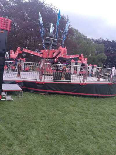 The image depicts an amusement park ride, known as "Evolution," in a grassy area, likely a fairground or park. The ride is red and black with bright lights. A metal safety fence surrounds the ride. Banners with the ride's name extend upwards. In the background, there are trees of varying colors, and the sky is overcast. There are a few blurred figures visible on the ride platform. The location is in Coventry, United Kingdom.