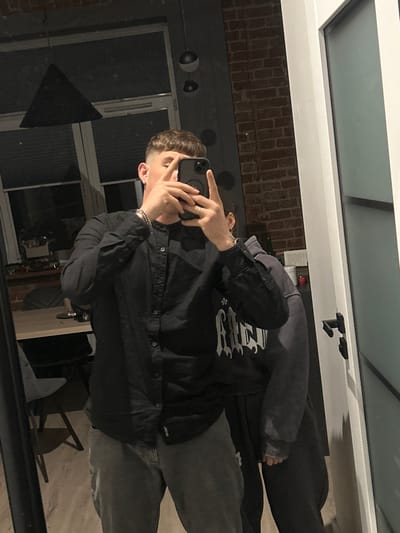 A man is taking a mirror selfie in what appears to be a dimly lit apartment. He is wearing a black button-up shirt and dark grey jeans. He has short, styled hair and is holding his phone with his right hand, obscuring his face. His left hand is raised, with his index finger extended. Another person, whose face is not visible, is standing behind him and to his right, wearing a grey hooded sweatshirt. The background shows a brick wall, a large window with blinds, a dining table with chairs, and a modern pendant light. The presence of a mirror suggests this is likely a residential interior, possibly a hallway or a living area. The image does not contain any elements that would place it specifically in the city of Siedlce, Poland.