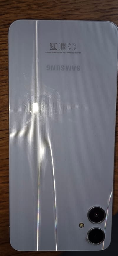 The image shows the back of a white Samsung smartphone resting on a wooden surface. The phone has a textured finish with vertical lines and features a dual-camera system on the lower right side. There are some scuff marks and smudges on the phone's surface, and a bright light reflection is visible in the upper left quadrant. Textual markings on the phone include "SAMSUNG" logo, "CE EAC TP BY", and "SM-A055F/DS Made in China IMEI:350988/75/65448/8". The background is a wooden surface with visible grain. There are no people or any other objects or activities depicted in the image. There is no indication of the location being in Marum, Netherlands.