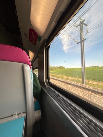 The image captures a unique, rotated perspective from inside a modern train carriage, looking out through a large window. The camera appears to be held in portrait orientation but depicts the horizontal exterior view at a tilt. Outside, a bright blue sky with scattered clouds dominates the upper right portion of the window, reflecting interior elements like lights and the cabin structure. Below the sky, lush green fields stretch into the distance, paralleled by train tracks that suggest the train's motion. A prominent electrical pylon and power lines are clearly reflected on the glass, superimposed over the outdoor landscape.
Inside the train, the cabin features a modern design. Part of a bright pink seat is visible in the foreground, leading to a grey and light blue seat. A passenger or an object covered by green fabric rests on the seat. Above the window, a white ceiling panel with an integrated light fixture is visible. A red oval sticker on the interior wall displays French text including "2.000 WIFI", "connectez-vous", and "sncf.com/appli", indicating this is an SNCF train offering Wi-Fi. The scene depicts a daytime journey through the sunny, rural French countryside near Essômes-sur-Marne, under clear weather conditions. The overall mood suggests a peaceful and comfortable travel experience.
