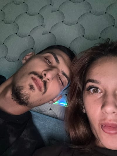 The image is a close-up, selfie-style photograph of two individuals. The person on the left, a male, is positioned at an angle, with his face tilted and one eye partially closed. He has dark hair, a mustache, and a goatee. The person on the right, a female, is looking directly at the camera with her mouth open and tongue slightly protruding. She has brown hair and is wearing an earring. They appear to be lying down or leaning against a surface with a textured, patterned background, possibly a wall or fabric. The lighting suggests an indoor setting. There is a hint of colorful light reflection on a surface between the two individuals. The overall impression is informal, possibly taken at night or in low light conditions. The location is not discernible from the image itself.