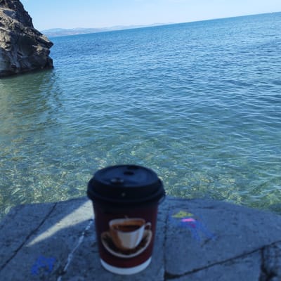 A disposable coffee cup with an image of a steaming cup of coffee is placed on a concrete surface. The cup is dark red with a white bottom and a black lid. The concrete surface is textured and has some faint blue and yellow chalk drawings on it. Behind the cup, a body of clear, turquoise water stretches out to the horizon under a bright blue sky. The water is calm with small ripples, and the rocky seabed is visible beneath the surface in the foreground. In the distance, a dark, rocky outcrop juts out from the left side of the frame into the water. Mountains are visible on the horizon in the far distance. The scene appears to be outdoors, likely at a seaside location, with the sun shining, suggesting it is daytime. The overall mood is serene and peaceful, evoking a sense of relaxation. No people are visible in the frame.