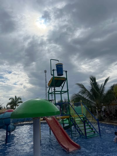 This image depicts an active aquatic recreational area within a water park located in Juwana, Indonesia. The scene is dominated by a large swimming pool with visible ripples on its blue surface. Above the pool, a tall, multi-level water play structure constructed from green and yellow metal tubing is present. This structure features a red water slide that descends into the pool and an accessible staircase with green steps and blue railings. At the apex of the structure, a large, blue tipping bucket, labeled "Kampoeng Goeroen Waterboom," is visible, with water actively flowing into it from an overhead pipe. In the foreground to the left, a green, mushroom-shaped water feature releases a continuous cascade of water into the pool. The sky is largely overcast with dense grey clouds, though the sun's bright disc is visible through a thinner patch of cloud cover in the upper center. To the far left in the background, a tall, grey telecommunication tower is visible. Multiple palm trees are scattered throughout the background, with a prominent one on the right displaying visible coconuts. Faint, indistinct figures appear to be seated under a shaded structure in the distant right background. A single individual is partially visible submerged in the water in the lower right corner of the frame.