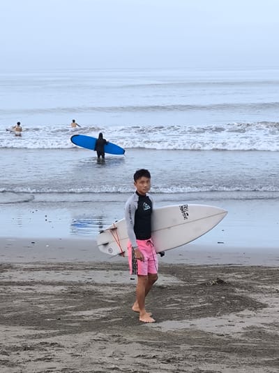The image depicts a man standing on a beach in Tibubeneng, Indonesia. He is holding a white surfboard under his right arm, wearing a black and grey rashguard and pink shorts. Behind him, other individuals are in the ocean, some with surfboards. The beach is dark sand, and the ocean waves are visible in the background under an overcast sky.