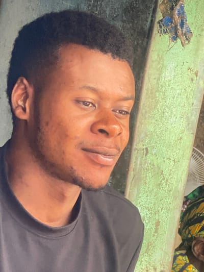 This image presents a close-up, head-and-shoulder portrait of a young male subject, of African descent, situated in Ibadan, Nigeria. The subject wears a dark, possibly black or charcoal grey, crew-neck t-shirt and has short, dark, textured hair with faded sides. His gaze is directed downwards and to the viewer's right, with a subtle, closed-mouth smile evident. Minor facial blemishes are present on the forehead and left cheek, and faint stubble outlines the jawline and chin.
The background to the immediate right of the subject is dominated by a worn, vertically oriented structure, likely a pillar or wall section, painted in a faded, mottled green color with visible texture and potential grime. An irregular, small piece of blue and brown patterned fabric is affixed to the upper section of this green structure. In the far right background, behind the green structure, a segment of a white electric fan's blade or grille is partially visible. Below the fan, a small portion of another person or object, clad in a multi-colored fabric with prominent yellow and green patterns, is discernible. The background behind the subject's left side and head is a plain, uniform, muted grey-green surface.