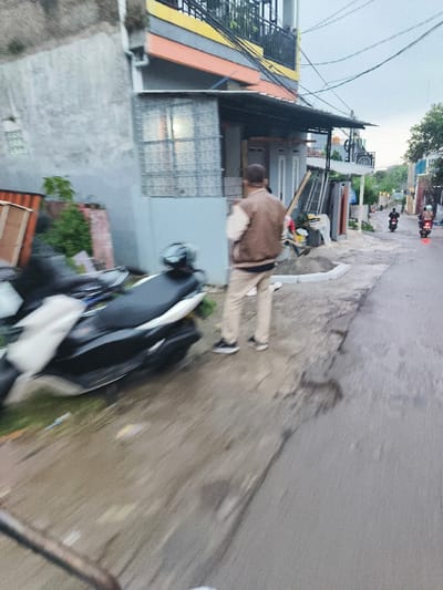 A man in a brown jacket and khaki pants is walking on a paved road in the city of Cimenyan, Indonesia, located at coordinates -6.887 latitude and 107.653 longitude. The road is lined by buildings on both sides, and there is a parked motorcycle to the left of the man. There are other vehicles, possibly motorcycles, traveling on the road in the distance, with a faint blur suggesting movement. The scene appears to be taken on a cloudy day. The man is walking away from the camera toward a building on the opposite side of the road. There is a partially visible, light gray, building on the left side of the frame. There is a small green plant growing next to the building. The road is slightly wet and appears to be in poor condition. The focus of the photo is on the man walking.