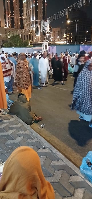 The image portrays a bustling nighttime street scene in Makkah Al Mukarramah, Saudi Arabia. In the foreground, a grey sidewalk with a chevron or herringbone paver pattern is visible. The top of a person's head and shoulder, covered by an orange hijab, occupies the lower left corner. To their right, at the edge of the asphalt street, an individual is seated or lying on the ground, enveloped in a dark olive or khaki-colored garment, with their bare feet exposed. A clear plastic water bottle lies discarded near this person. On the far right foreground, a translucent blue plastic bag containing small, dark, granular objects is present on the pavement.
The mid-ground features a dense crowd of pedestrians, primarily adults, walking along the street. Individuals are dressed in a variety of attire; several men wear traditional white thobes and head coverings, while others wear darker robes or lighter tunics. Women are observed wearing hijabs, niqabs, or patterned modest clothing. The background is dominated by numerous tall, modern high-rise buildings with illuminated windows and facades. Decorative string lights with individual bulbs are strung horizontally across the street overhead. A prominent utility pole or cell tower with multiple antennas is centrally located in the mid-background. Construction barriers or temporary partitions are also visible behind the crowd.