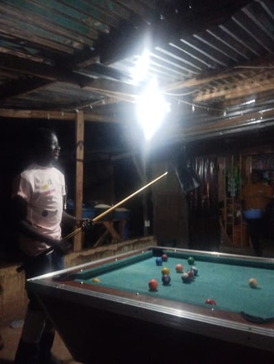 In a dimly lit, corrugated-roofed space, a person in a light-colored shirt stands beside a pool table, cue stick in hand. The table is arranged with several colorful billiard balls. Overhead, a bright, single light fixture illuminates the scene, casting stark shadows. The setting appears to be a casual, possibly outdoor, recreation area. Other individuals are visible in the background.
